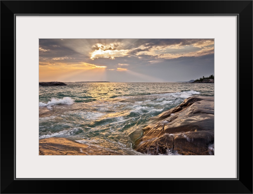 Burst of sun rays peeking out behind a cloud as waves lap on to a rocky shore.