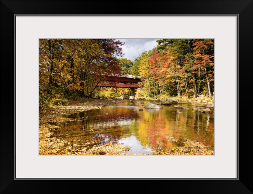 A photograph of a covered bridge spanning a stream in a forest in autumn foliage.