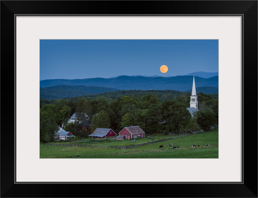 A photograph of a small village in the countryside seen at night under a clear sky and a full moon.