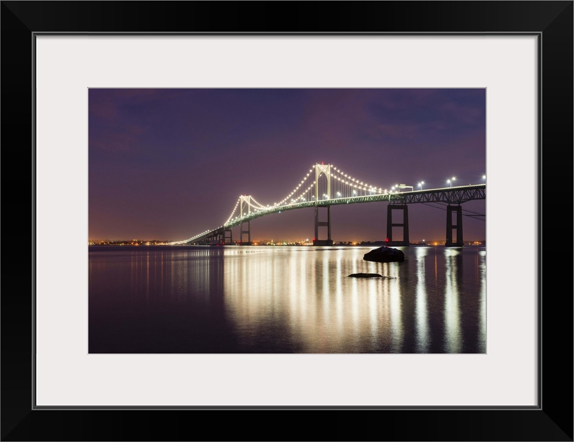 A photograph of a large suspension bridge seen lit up at night casting long reflections on the water below.