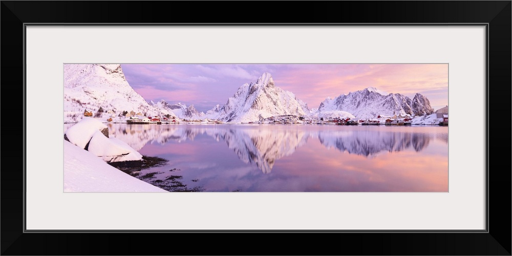 A photograph of a snow covered mountain range in winter casting a clear reflection in the fjord below.