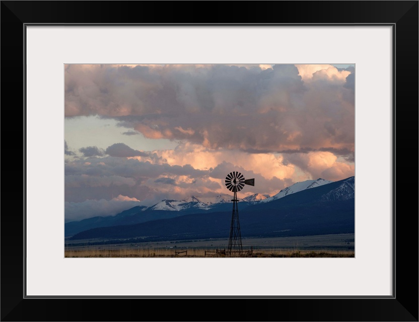 Landscape photograph of a windmill in a field with snow covered mountain peaks in the distance.