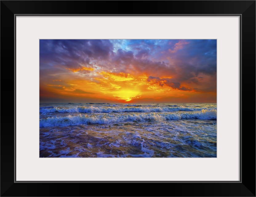 Purple and blue clouds hover above a brilliant red and orange sunset. A cold blue ocean with sunset illuminated breaking w...