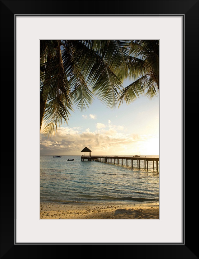 Vertical photograph of a tropical landscape framed with palm trees on both sides and a long pier leading to the ocean in t...