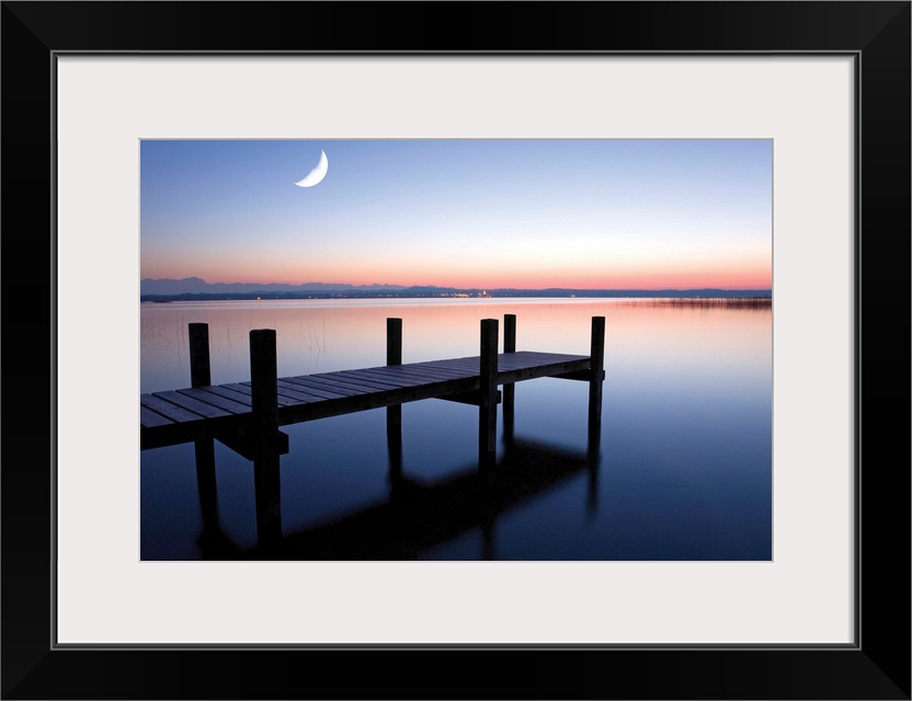 Sunset photograph of a dock over calm water and a half moon in the sky.