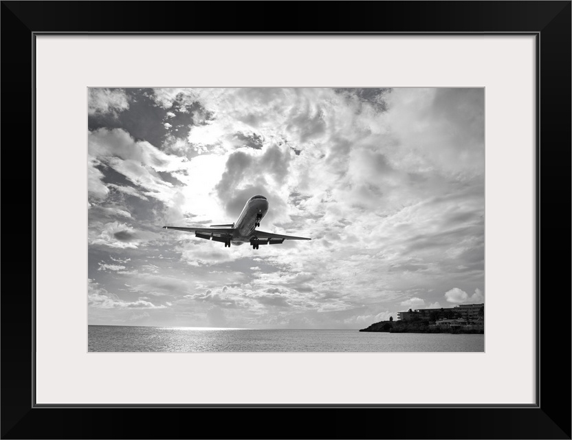 Black and white photograph taken of a commercial plane descending over ocean water about to land on an island.