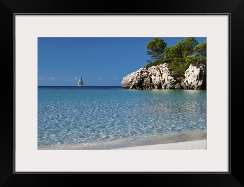 Photo of crystal clear blue water and white sand beach in Spain with a sailboat in the distance and a large cliff jutting ...