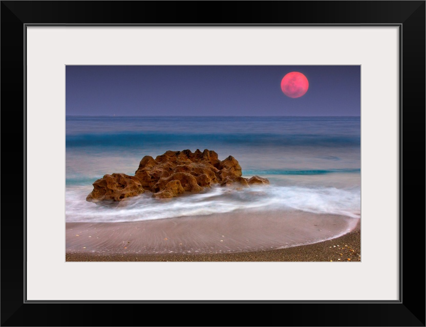 A unique rock formation sits in the ocean water which is photographed under a deep red moon.