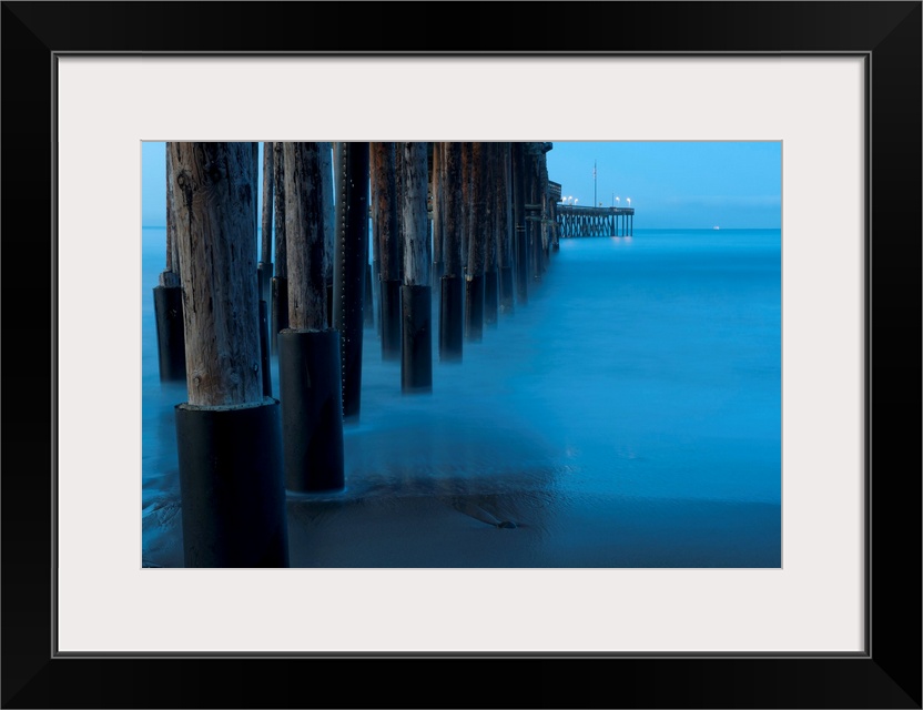 Large print of tall wooden pier pillars holding up a pier leading into the ocean from the shore.