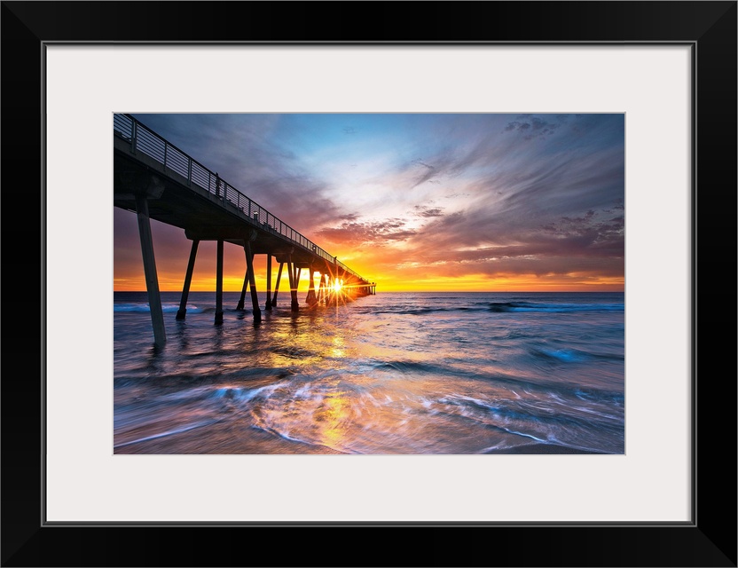 Sun intersects pier at sunset with colorful waves and sky.