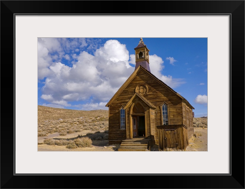 Large, horizontal photograph of a small, old wooden church surrounded by empty dessert, beneath a blue sky with fluffy clo...