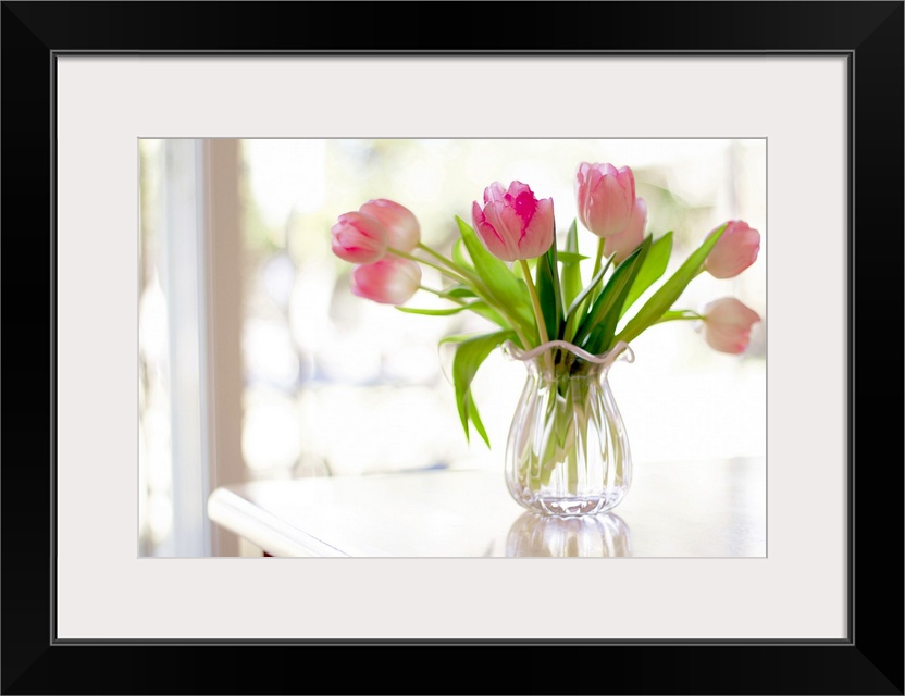 Ruffled pink glass vase filled with soft, pink tulips sitting on table in front of window bright with sunlight.