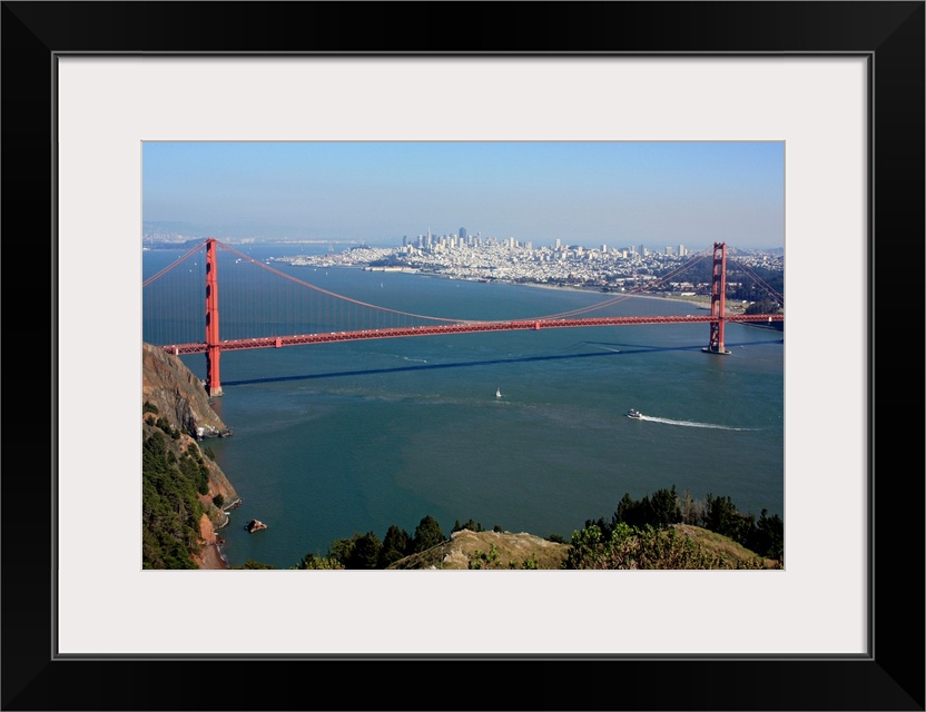 This is an aerial landscape photograph of the suspension bridge, bay, and distant city skyline on a bright sunny day.