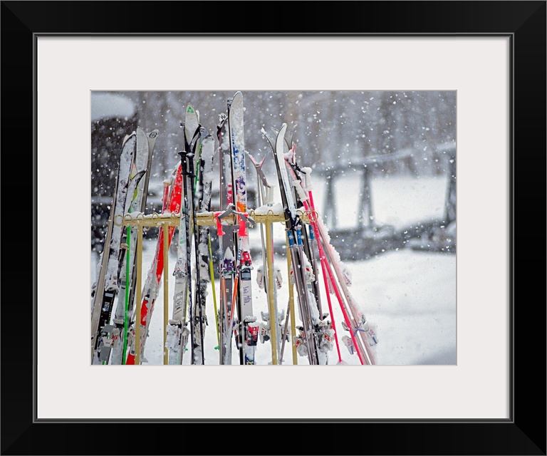 In this photograph multiple skis are stored on an outside rack as large white snowflakes fall all around. Blurred backgrou...