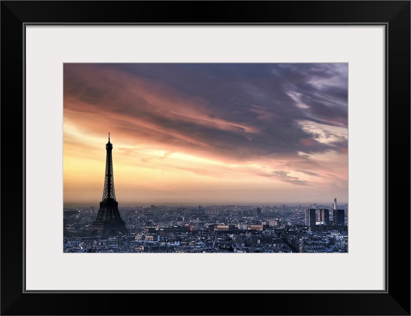 Big, horizontal photograph of the sun rising through wispy clouds as the Eiffel Tower looms over the city of Paris, France.