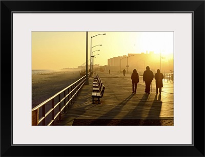 Three people walking down boardwalk near beach at sunset.
