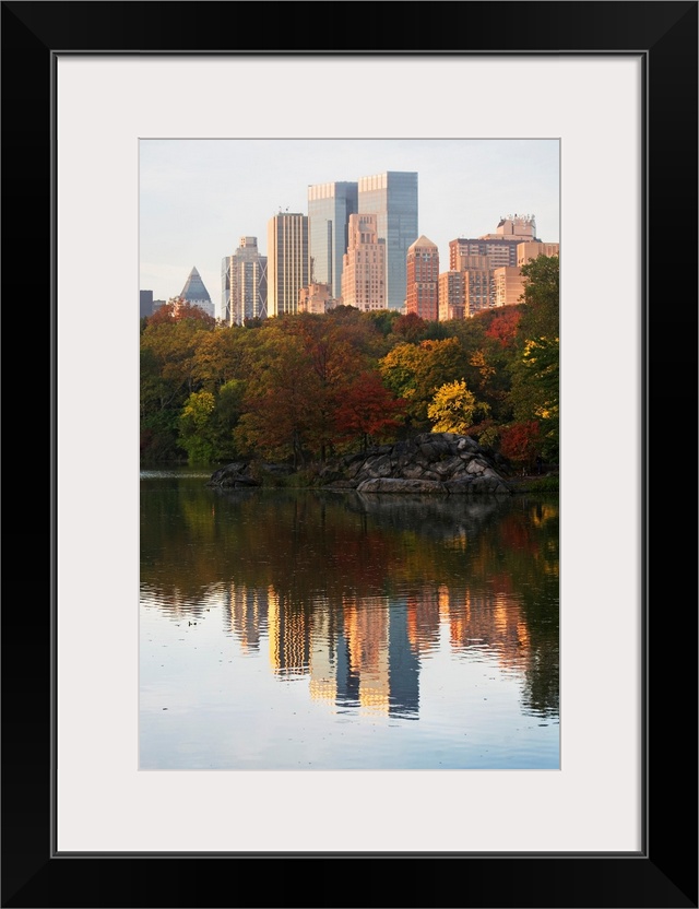 This is a vertical photograph of autumn trees and skyscrapers reflecting in the calm waters of this city park pond.