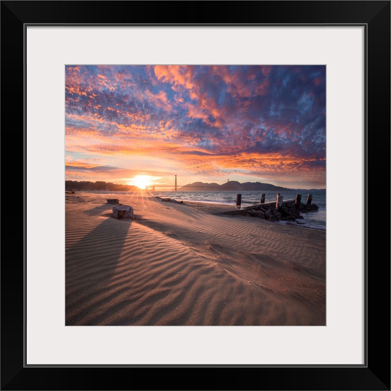A square photograph of a beach with the golden gate bridge in the background at sunset.