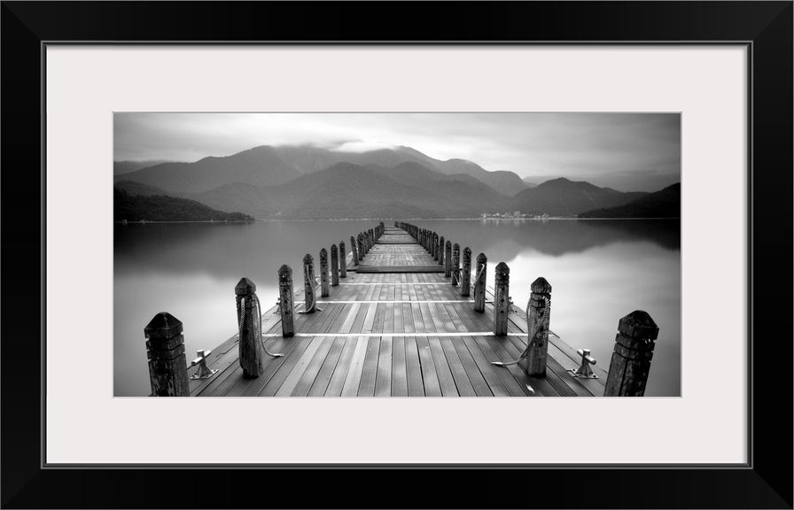 A black and white photograph of a long pier at a lake with large mountains in the distance.