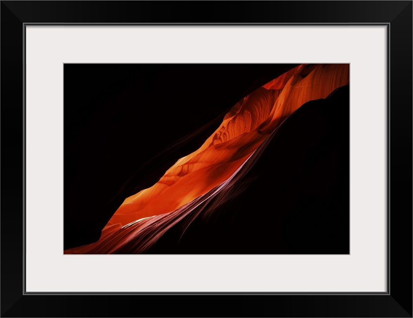 Large, landscape photograph of darkness surrounding a diagonal, thin area of a sandstone formation in Antelope Canyon, in ...