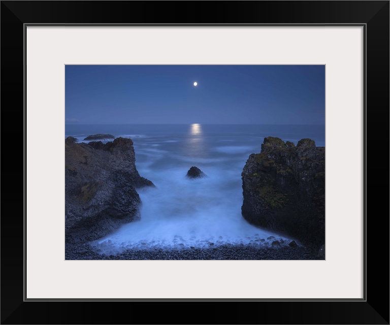 Illuminated moon in the distance hanging over the ocean in the distance, with two bookend boulders in the foreground.