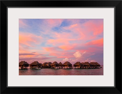 A vibrant pink and red sunset over bungalows on stilts over the water