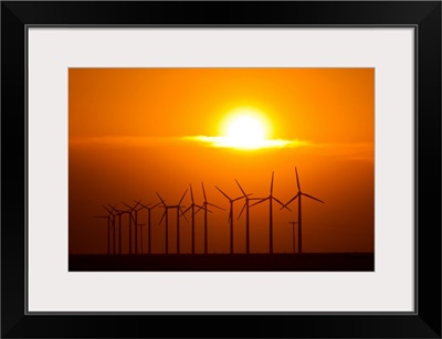The sun sets behind a row of spinning windmills or wind turbines