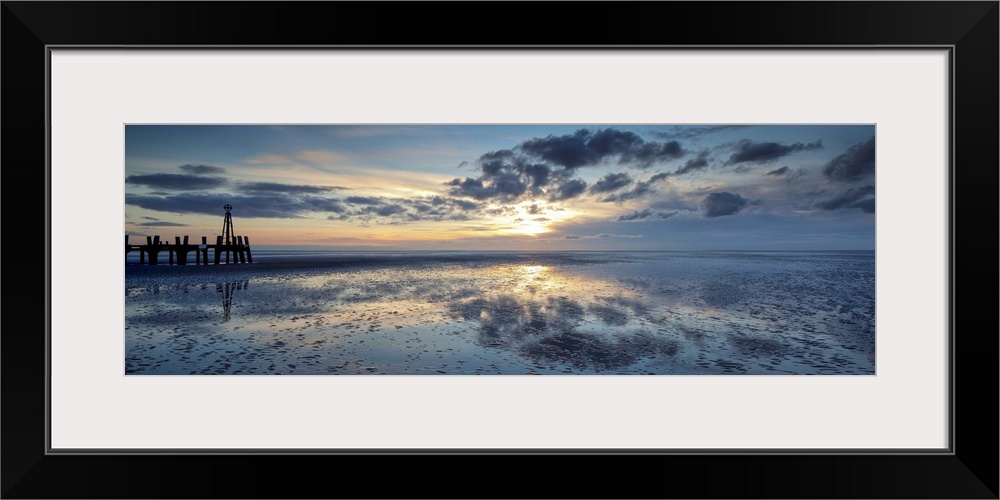 A panoramic image of a tranquil golden blue dawn with fluffy clouds over reflecting wet sand with a harbour installation.