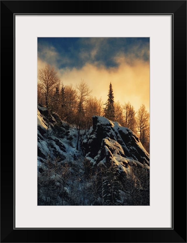 Clouds lit up with fading sunlight over a forest near snowy rocks on a mountainside.