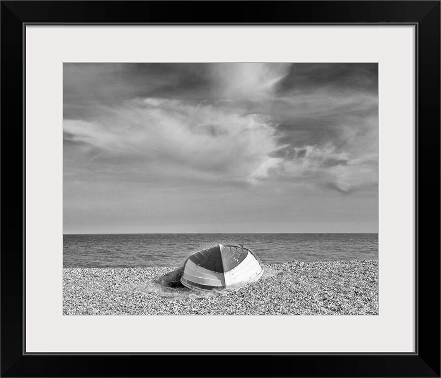 A monochrome black and white image of an upturned rowing boat on an English shingle beach in bright sunshine with fluffy w...