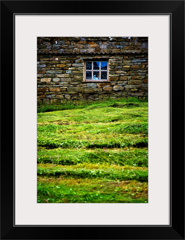 Photograph taken of a stone wall with a window in it and thick cut grass just in front.