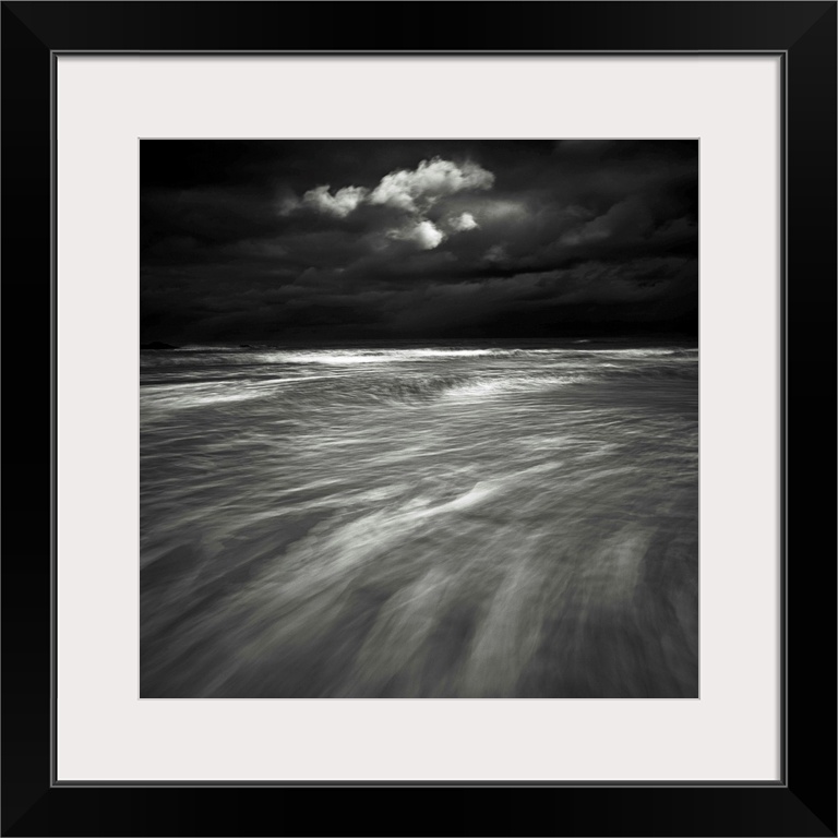 A monochrome black and white sepia toned dramatic image of swooshing wavws and a lone cloud in a dark stormy sky over a se...