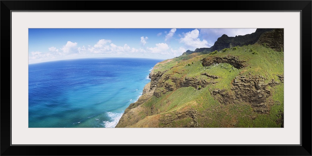 Panoramic photograph of rocky grass covered cliff near water's edge under a cloudy sky.