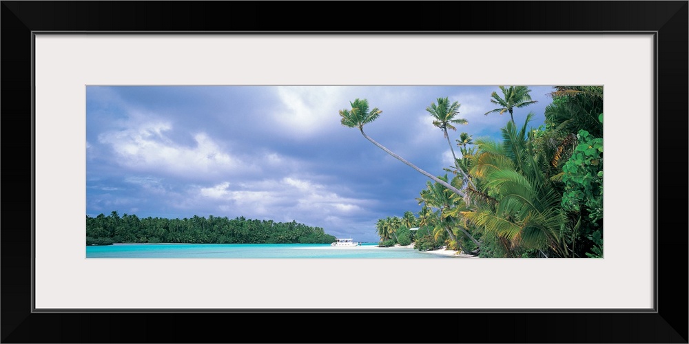 Panoramic photograph of lagoon with shoreline full of trees and tree covered island in distance under a cloudy sky.