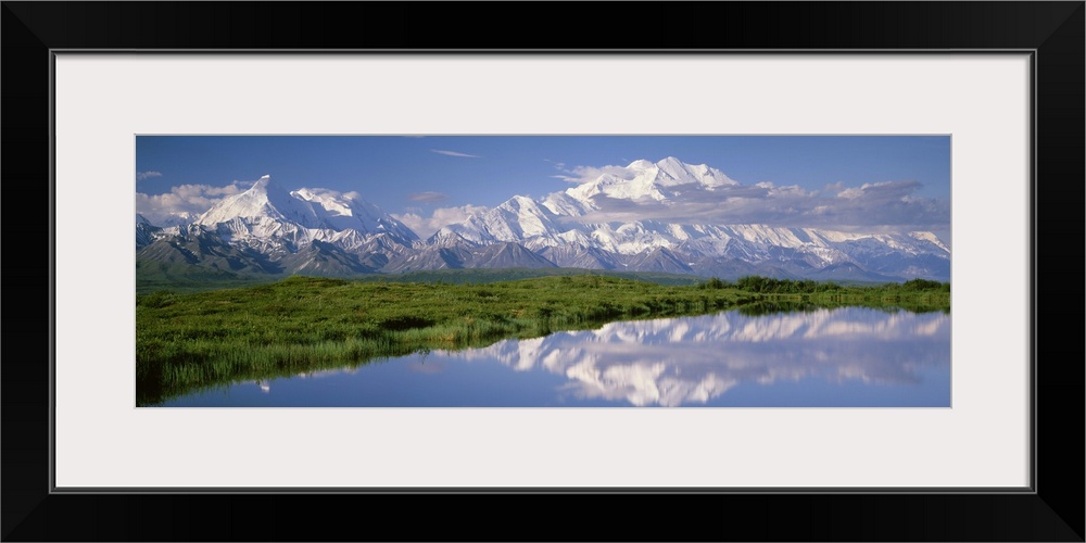 A snow covered mountain range is photographed in panoramic view with a large field shown just in front and a body of water...