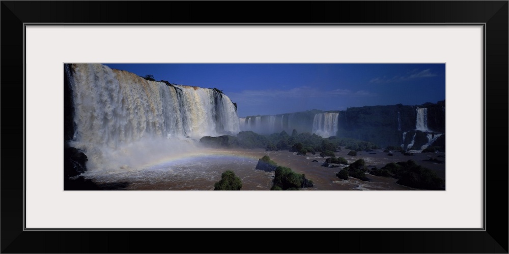 This is a panoramic photograph of enormous South American waterfalls and a rainbow forming under the cascades.