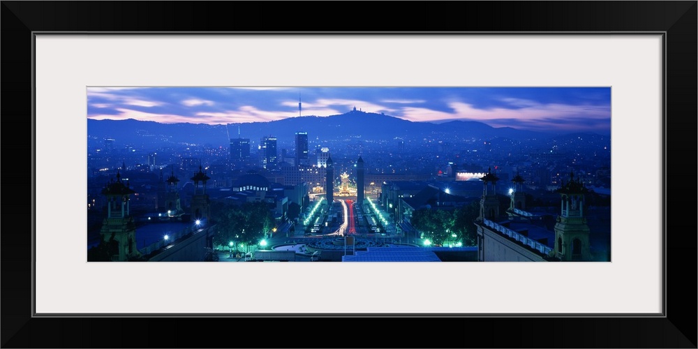 Giant, high angle photograph of Barcelona, lit at night, mountains in the background beneath a mostly cloudy sky, in Spain.