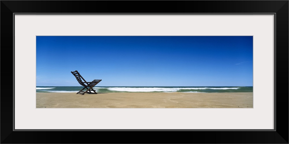 Huge panoramic piece of a lone beach chair skewed to the left sitting in the sand with waves crashing and a clear blue sky...