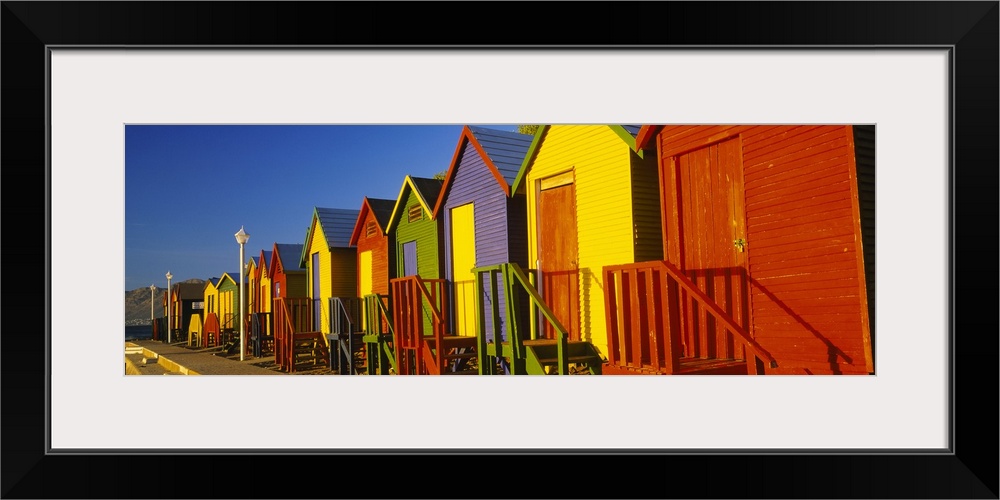 Panoramic photograph displays a line of vibrantly colored seaside shacks on a sunny day.  In the background there is a mou...