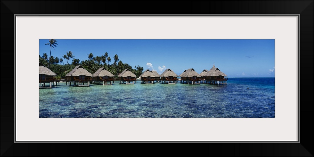 Panoramic photograph showcases a row of small shacks sitting in the middle of clear water from the Pacific Ocean.  Located...