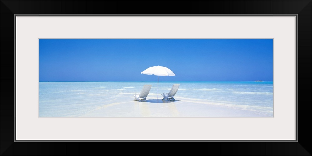 A panoramic photograph displaying two chairs sitting underneath an umbrella on a sandy beach in Maldives.  The clear water...