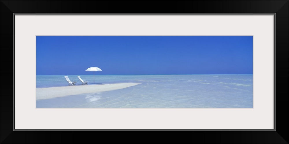 Large panoramic picture of two white chairs and a white beach umbrella sitting on sand that stretches into the crystal cle...