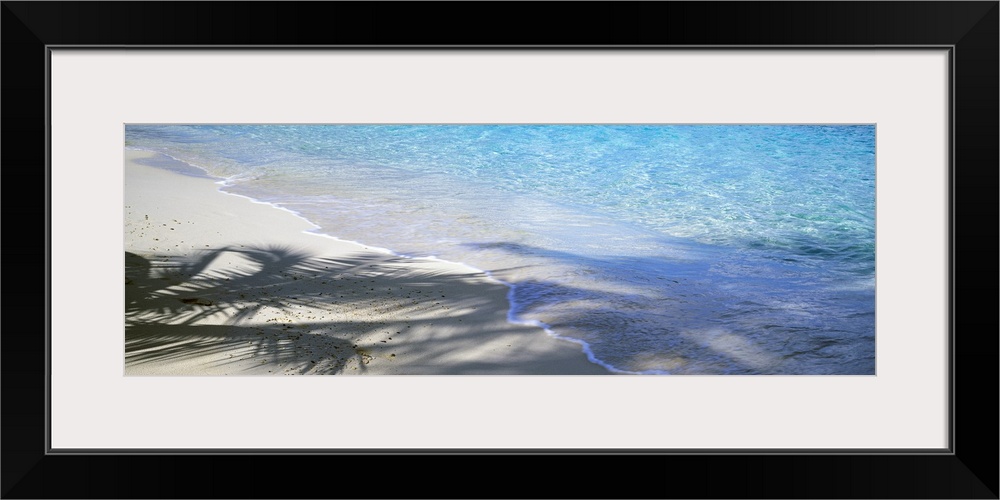 Panoramic shot of a beach with crystal clear water rushing up onto the sand that is partially shadowed by palm trees.