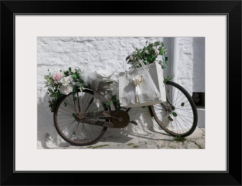 A bike with rusty gears covered in beautiful roses with a decorative ribboned tote bag leaning against a textured wall.