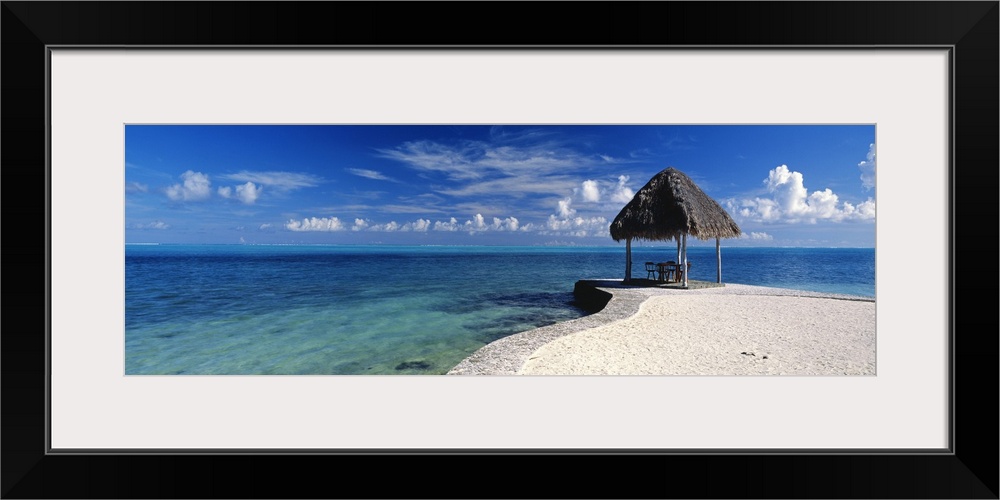 Panoramic view of a small straw hut sits on the beach that protrudes out into the clear blue ocean.