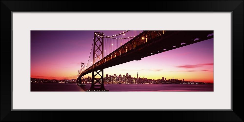 Large panoramic photo print of a long bridge leading to a lit up city off in the distance at sunset.