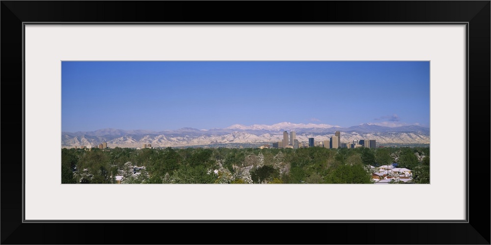 The city of Denver is photographed from a distance over trees and homes. Mountains are pictured far off in the background.