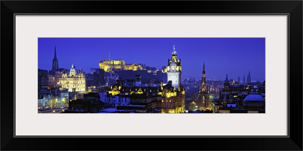 Panoramic photograph taken of buildings in Scotland lit up at night with a castle shown in the background.