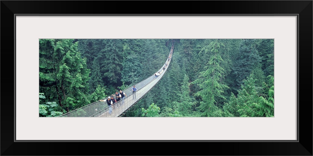 Long horizontal photo on canvas of people walking across a long bridge that runs through the top of a forest.