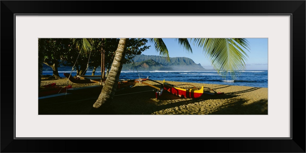 A large panoramic photograph of a boat on the beach that is mostly covered by the shadows of palm trees. Water and mountai...